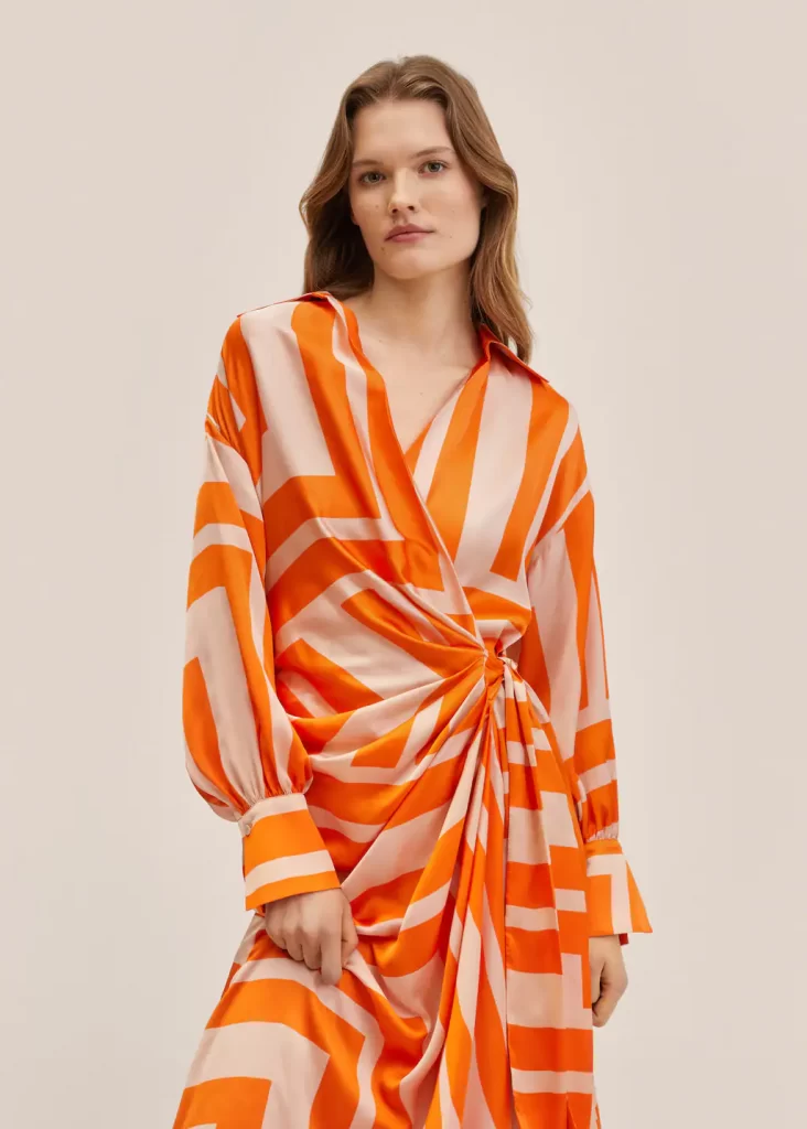 Graphic printed orange and beige satin midi dress with twisted knot at waist and slit at leg. Open v neck with collar and long sleeves with cuffs. 