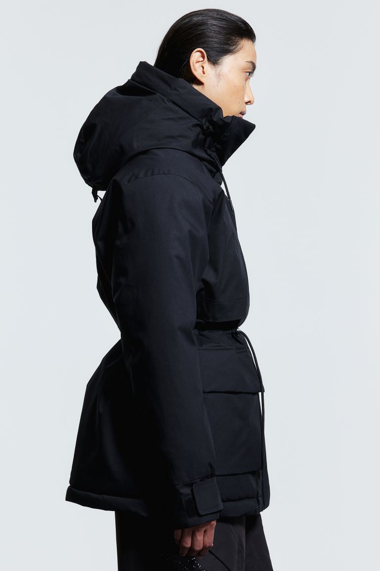 H&M Two-layer black parka in windproof, water-repellent functional fabric designed to keep you dry and warm during light showers and protect you from the wind. Regular fit with a hood that has a drawstring with cord stoppers at the front and an adjustable grosgrain tab with a buckle at the back to help it stay in place. Zip down the front with an anti-chafe chin guard and a wind flap with concealed press-studs. Long sleeves with a hook and loop fastener at the cuffs and ribbed inner cuffs. Zipped chest pockets, front flap pockets with concealed press-studs, and an inner pocket. Concealed drawstring with cord stoppers at the waist and hem for an adjustable fit. Lined. Breathability 10,000 g/m2/24 h. Designed with THERMOLITE® EcoMade T-Down filling, an alternative to down but with higher loft, better moisture resistance and high air permeability. It is also warmer when wet and dries faster.